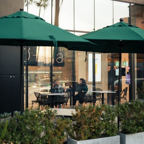 Two people seated at a table inside Olia Restaurant, viewed through a window with green umbrellas.
