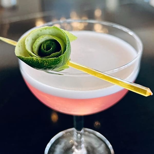 A pink cocktail with a white foam top, garnished with a lime peel rose on a yellow pick.