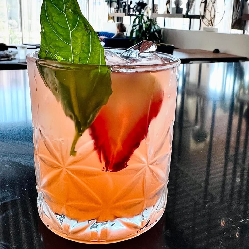 A pink cocktail with a large basil leaf and ice, on a dark bar top.