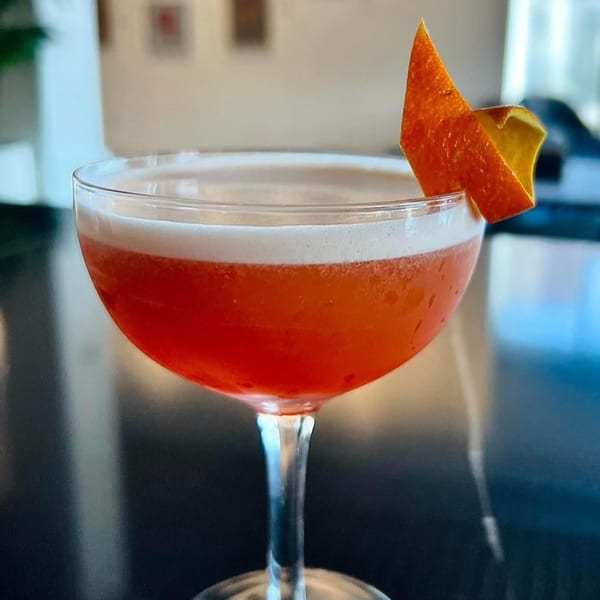 A vibrant red cocktail with a foamy top, garnished with an orange peel, on a dark surface.