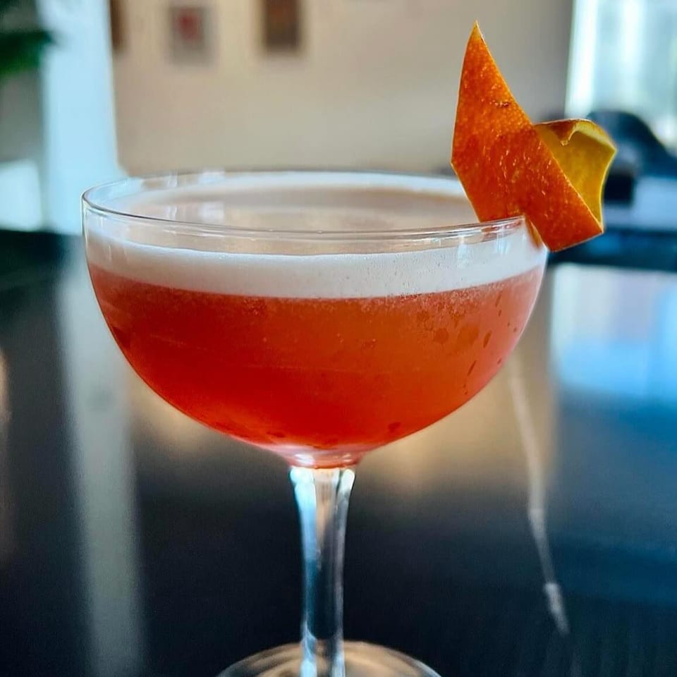 A vibrant red cocktail with a foamy top, garnished with an orange peel, on a dark surface.