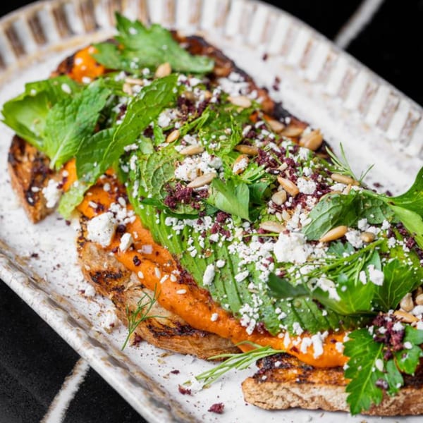 Mediterranean toast with red spread, avocado, feta, herbs, and pine nuts on a white plate.