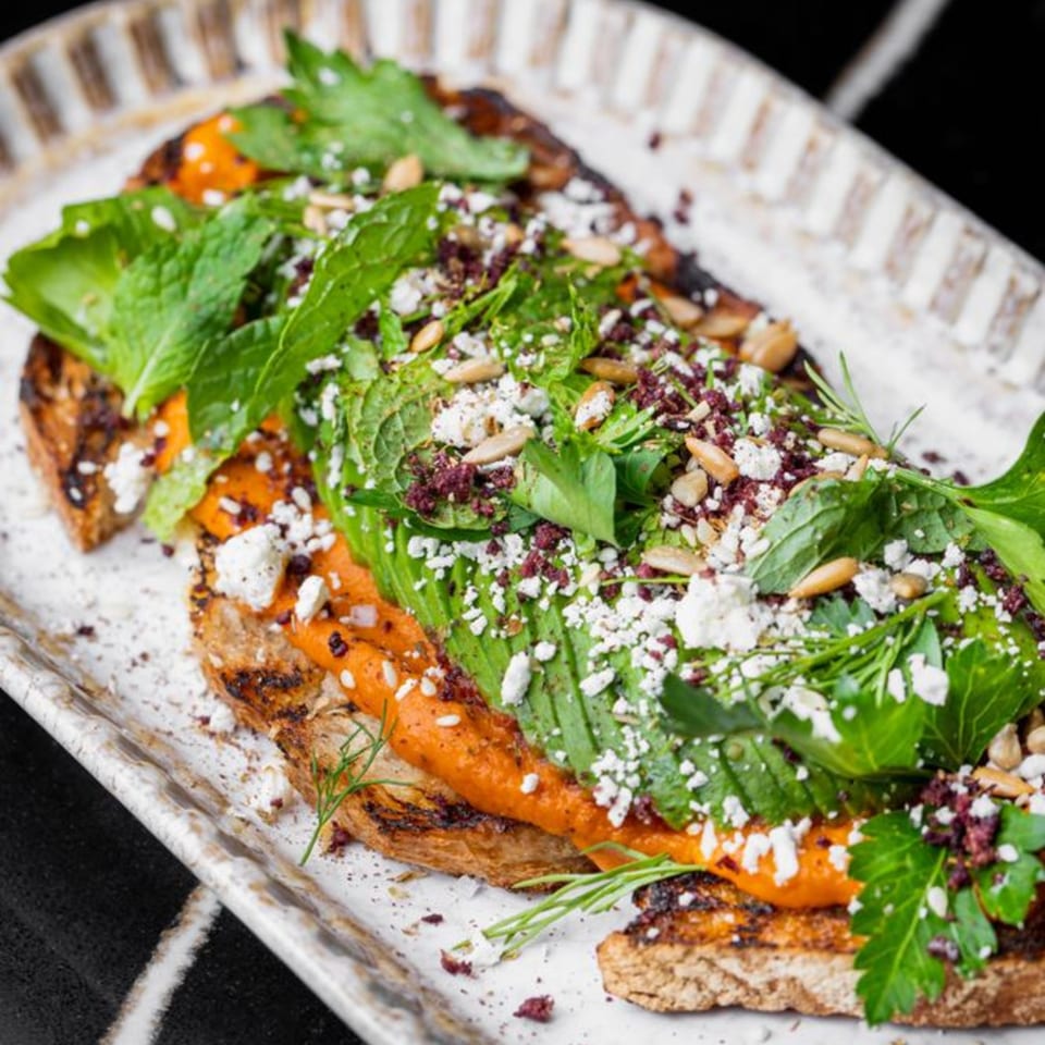 Mediterranean toast with red spread, avocado, feta, herbs, and pine nuts on a white plate.