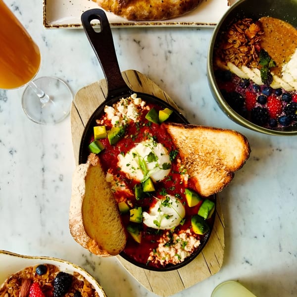 Shakshuka with poached eggs, avocado, feta, and toast, alongside a fruit and granola bowl.