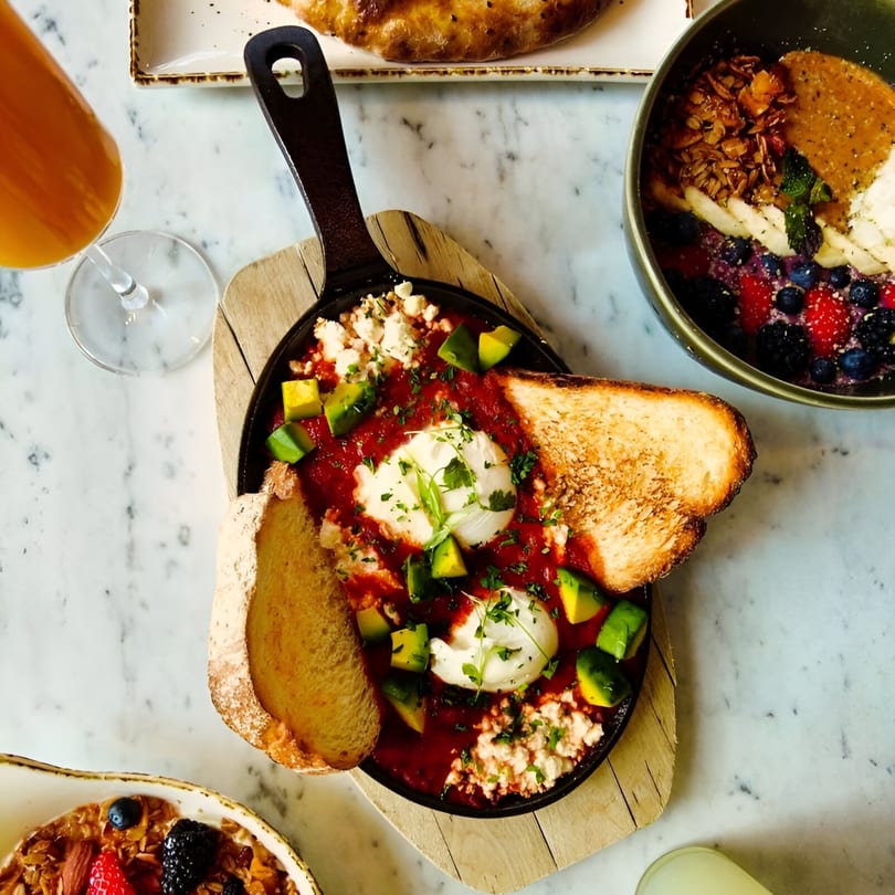 Shakshuka with poached eggs, avocado, feta, and toast, alongside a fruit and granola bowl.